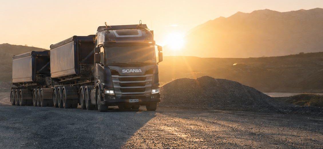 A dark blue semi-truck with trailers parked on the gravel lot of an earthworks site at sunset